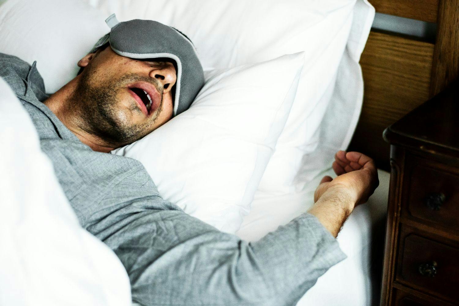 man in bed with a sleep mask on and snoring with his mouth open.