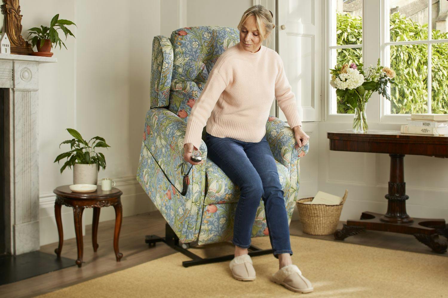 Woman exiting her chair in the rise position with a Morris & Co. Golden Lily print Oak Tree Mobility rise and recline chair in a living room. 