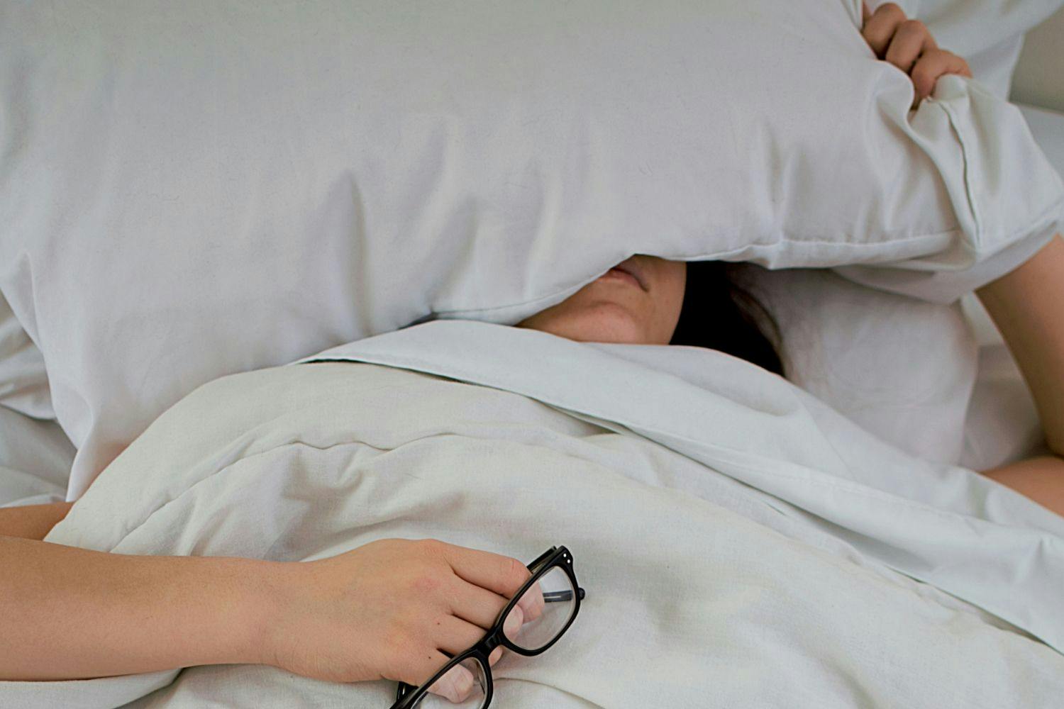 woman in bed with a pillow over her eyes and holding a pair of glasses