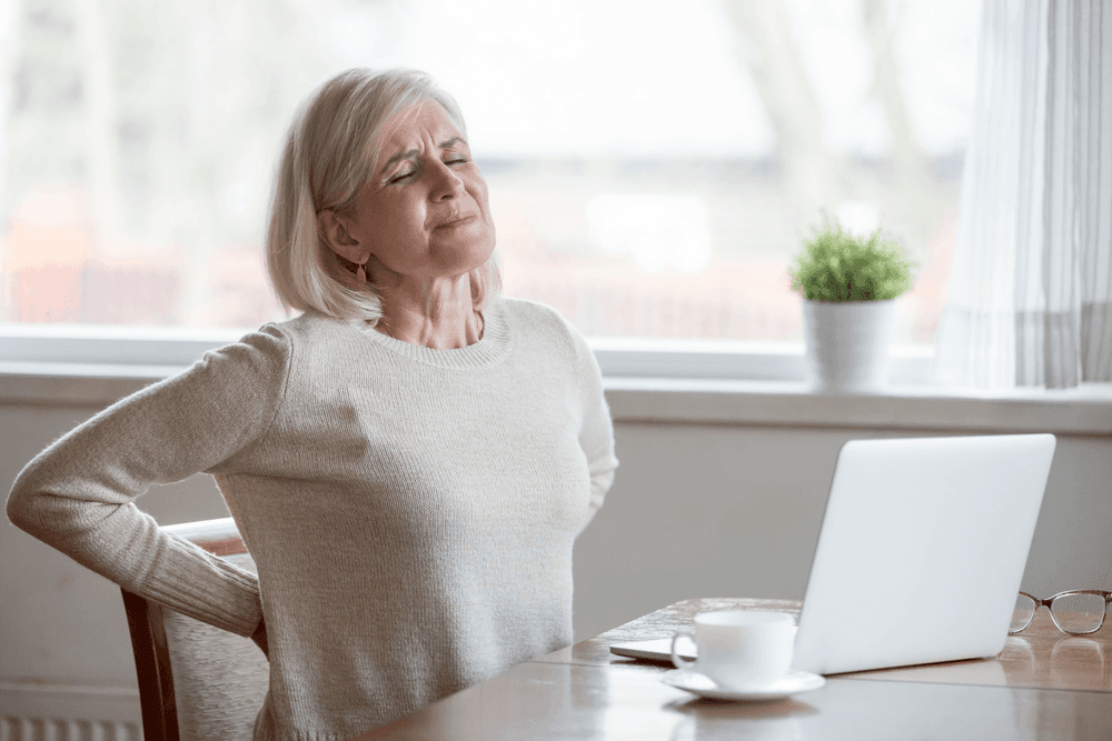Woman sat at desk with her laptop open. She is arching her back in pain due to her posture