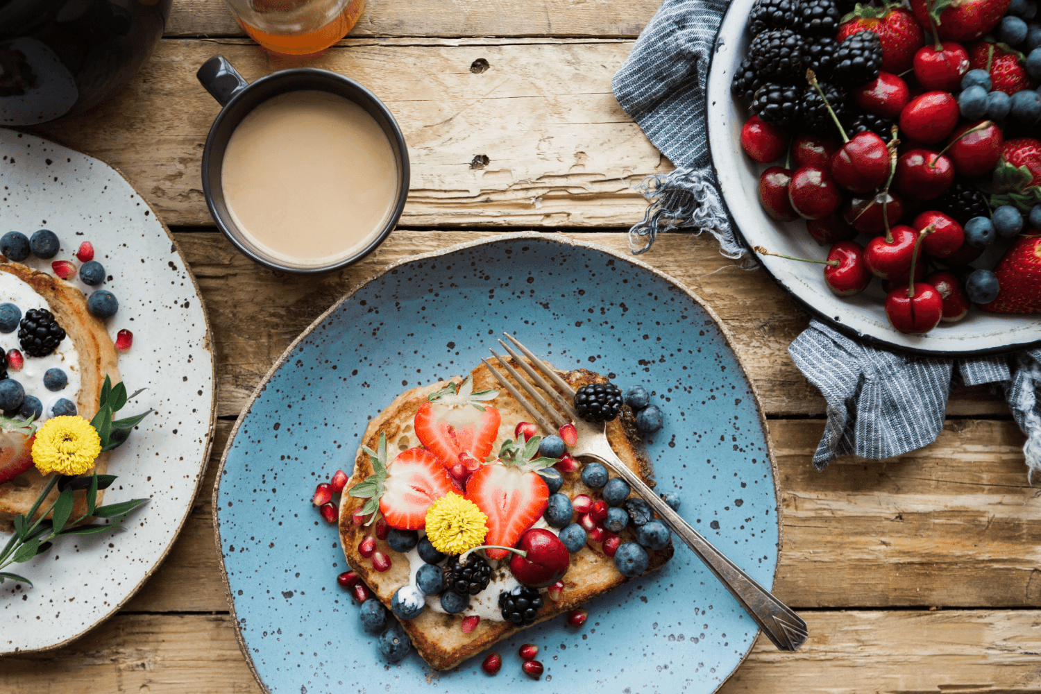 wooden table with cup of tea, blue plates, bowl with berries and toast with berries on top.