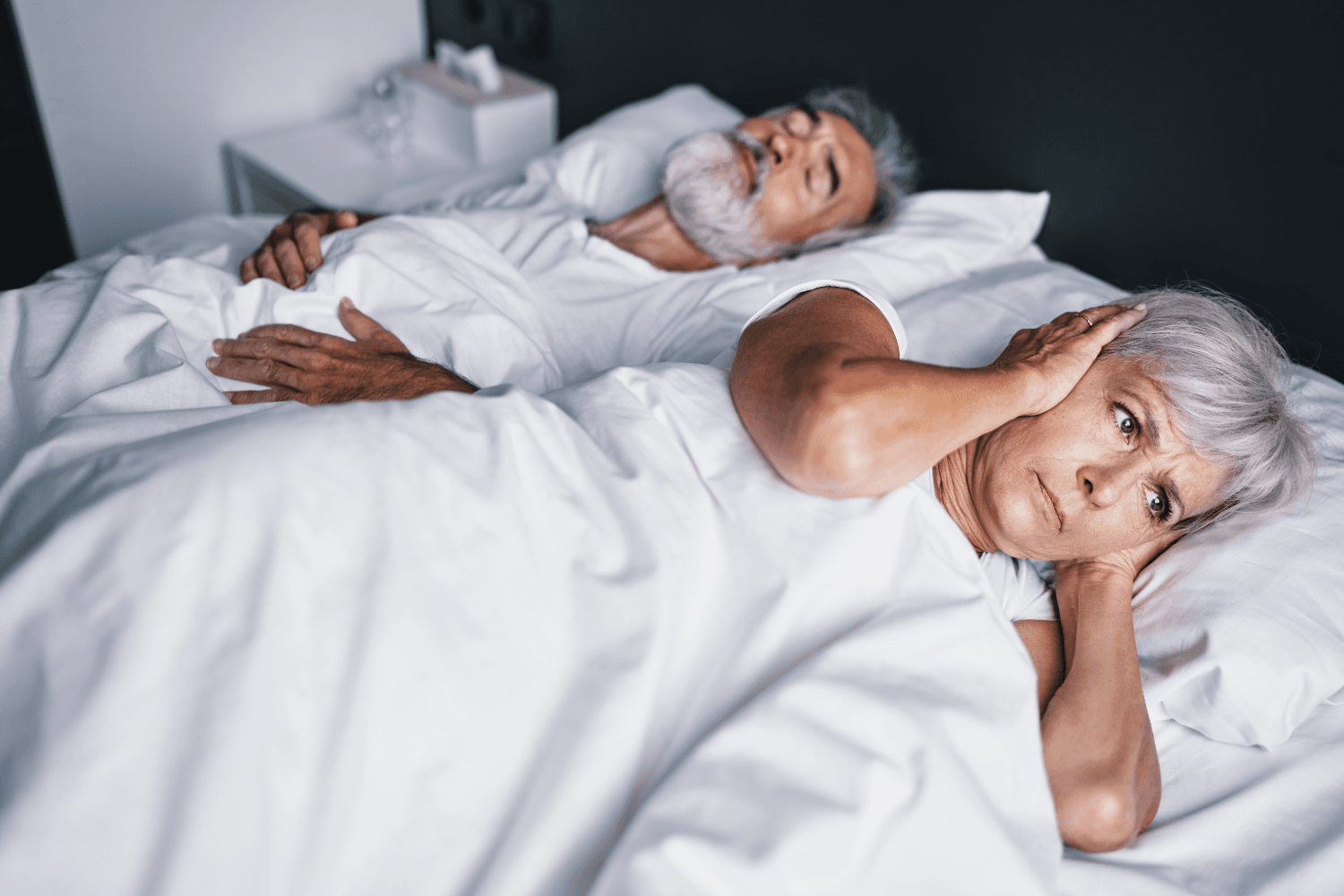 man and woman lying in bed sleeping, woman covering ears as man snores