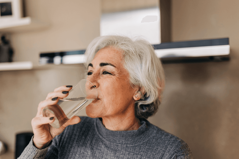 Older woman in a grey polo jumper smiling as she drinks a glass of water in the kitchen