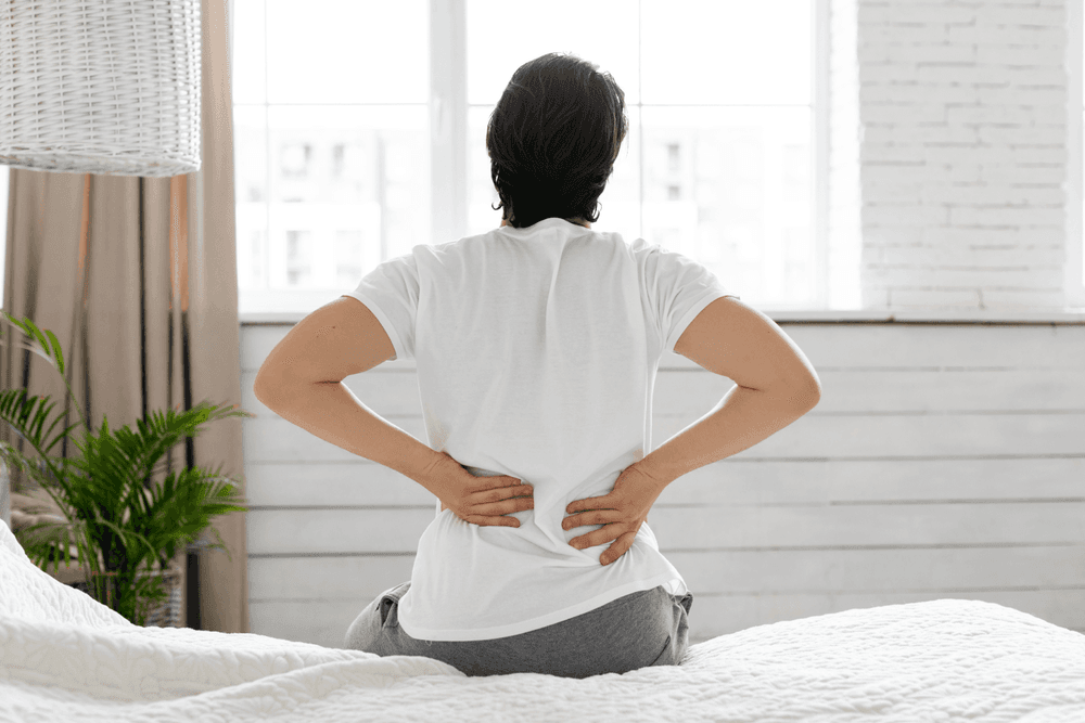 Rear view of a man holding his lower back in discomfort whilst sitting on his bed